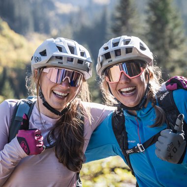 Zwei lachende Frauen mit Fahrradhelmen und Sonnenbrillen, die in einer grünen Berglandschaft stehen und Daumen hoch zeigen. | © Kleinwalsertal Tourismus eGen • Fotograf: Bastian Morell
