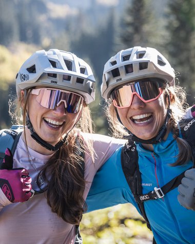 Zwei lachende Frauen mit Fahrradhelmen und Sonnenbrillen, die in einer grünen Berglandschaft stehen und Daumen hoch zeigen. | © Kleinwalsertal Tourismus eGen • Fotograf: Bastian Morell