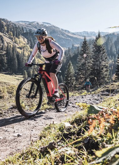 Frau fährt mountainbike auf einer schmalen Waldweg im Bärgunttal, umgeben von Bergen und Tannenbäumen, sonniges Wetter. | © Kleinwalsertal Tourismus eGen • Fotograf: Bastian Morell