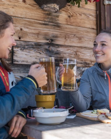 Zwei Frauen sitzen im Freien vor einer Holzhütte, trinken Bier und lachen. Sie haben Essen auf dem Tisch vor sich. | © Kleinwalsertal Tourismus eGen • Fotograf: Bastian Morell