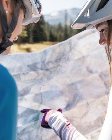 Zwei Mountainbikerinnen schauen auf einen Stadtplan während einer Bike-Tour in den Bergen, beide tragen Helme und Fahrradkleidung. | © Kleinwalsertal Tourismus eGen • Fotograf: Bastian Morell