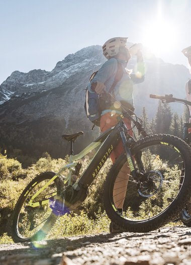 Zwei Mountainbikerinnen am Berg mit Sonnenlicht im Hintergrund, umgeben von Natur und Bergen. | © Kleinwalsertal Tourismus eGen • Fotograf: Bastian Morell