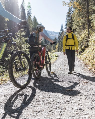 Zwei Mountainbiker und eine wandernde Person auf einem schottrigen Waldweg in Bärgunttal, umgeben von Bäumen an einem sonnigen Tag. | © Kleinwalsertal Tourismus eGen • Fotograf: Bastian Morell