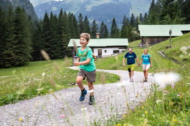 Familie beim Wandern im Gemsteltal, ein Junge läuft lachend vor der Gruppe, umgeben von grüner Natur und Bergen. | © Kleinwalsertal Tourismus eGen • Fotograf: Bastian Morell