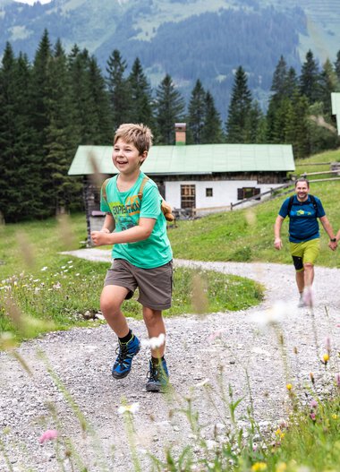 Familie beim Wandern im Gemsteltal, ein Junge läuft lachend vor der Gruppe, umgeben von grüner Natur und Bergen. | © Kleinwalsertal Tourismus eGen • Fotograf: Bastian Morell