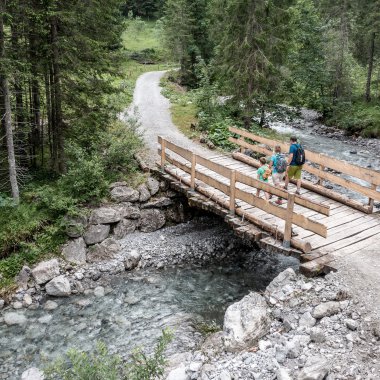 Familie beim Wandern auf einer Holzbrücke in einem Waldgebiet im Gemsteltal, umgeben von Bäumen und einem Fluss unter der Brücke. | © Kleinwalsertal Tourismus eGen • Fotograf: Bastian Morell