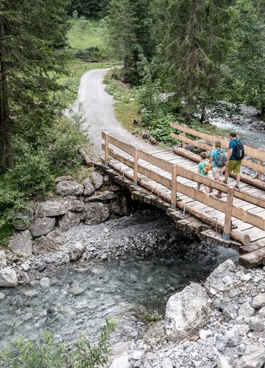 Familie beim Wandern auf einer Holzbrücke in einem Waldgebiet im Gemsteltal, umgeben von Bäumen und einem Fluss unter der Brücke. | © Kleinwalsertal Tourismus eGen • Fotograf: Bastian Morell