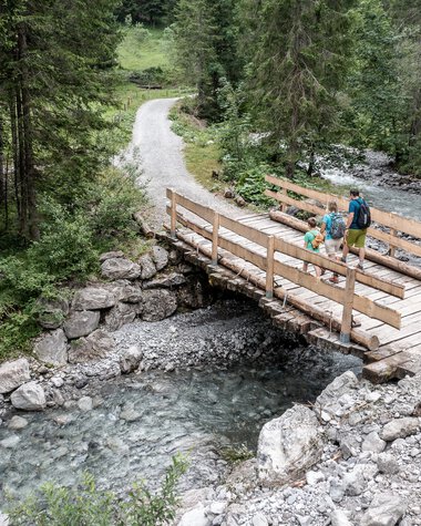 Familie beim Wandern auf einer Holzbrücke in einem Waldgebiet im Gemsteltal, umgeben von Bäumen und einem Fluss unter der Brücke. | © Kleinwalsertal Tourismus eGen • Fotograf: Bastian Morell