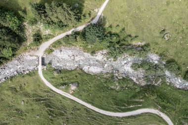 Luftaufnahme eines Wanderwegs entlang eines Flusses in einer grünen Landschaft, umgeben von Bäumen und Wiesen. | © Kleinwalsertal Tourismus eGen • Fotograf: Bastian Morell