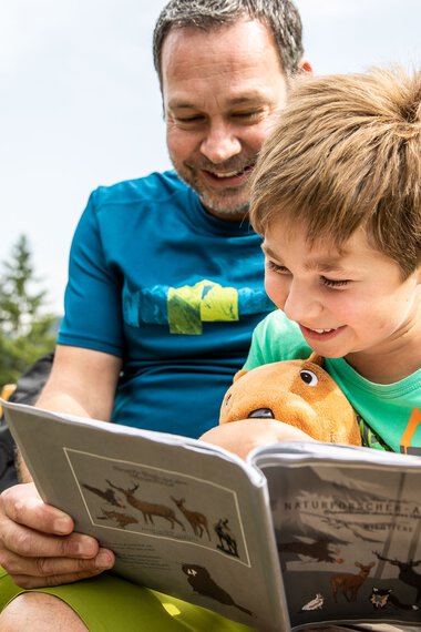 Vater und Sohn sitzen im Grünen und lesen gemeinsam ein Bilderbuch in einer Berglandschaft. | © Kleinwalsertal Tourismus eGen • Fotograf: Bastian Morell