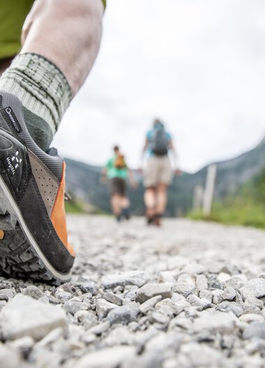 Close-up of a hiker's foot in a mountain shoe on a rocky trail in the mountains with a group of hikers in the background in nature. | © Kleinwalsertal Tourismus eGen • Photographer: Bastian Morell