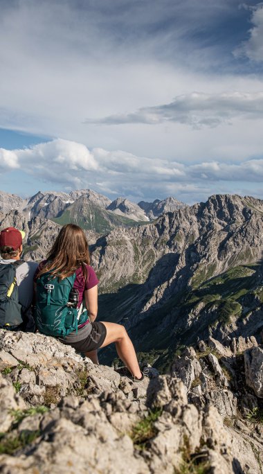 Zwei Wanderer mit Rucksäcken sitzen auf einem Felsen und genießen die Aussicht auf die Berge bei einer Wanderung auf der Kanzelwand. | © Kleinwalsertal Tourismus eGen • Fotograf: Bastian Morell