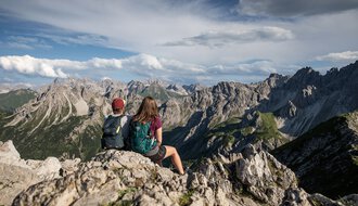 Zwei Wanderer mit Rucksäcken sitzen auf einem Felsen und genießen die Aussicht auf die Berge bei einer Wanderung auf der Kanzelwand. | © Kleinwalsertal Tourismus eGen • Fotograf: Bastian Morell