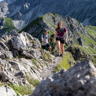 Zwei Wanderer auf einem schmalen Bergpfad mit Felsen und grünen Berghängen während einer Wanderung in den Alpen. | © Kleinwalsertal Tourismus eGen • Fotograf: Bastian Morell