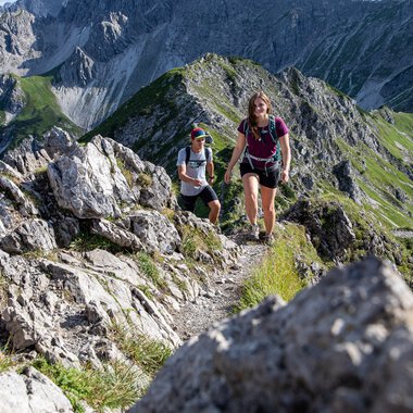 Zwei Wanderer auf einem schmalen Bergpfad mit Felsen und grünen Berghängen während einer Wanderung in den Alpen. | © Kleinwalsertal Tourismus eGen • Fotograf: Bastian Morell