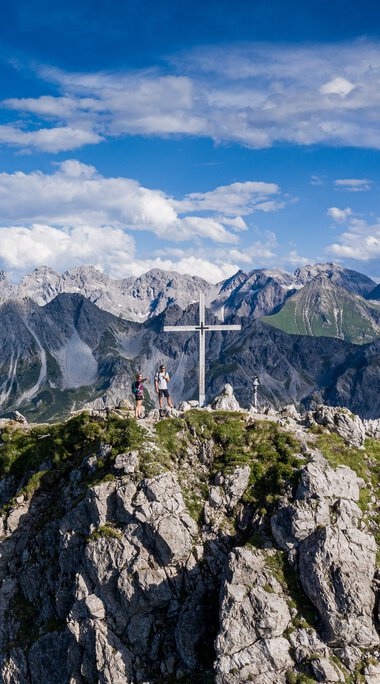 Berggipfel mit Gipfelkreuz und Wanderern, oberhalb der Alpen, bei schönem Wetter, mit blauer Himmel und Wolken. | © Kleinwalsertal Tourismus eGen • Fotograf: Bastian Morell