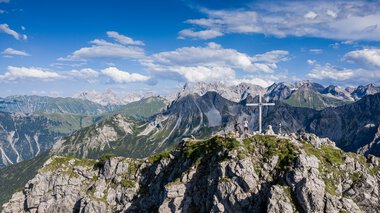 © Kleinwalsertal Tourismus eGen • Fotograf: Bastian Morell Berggipfel mit Gipfelkreuz und Wanderern, oberhalb der Alpen, bei schönem Wetter, mit blauer Himmel und Wolken. | © Kleinwalsertal Tourismus eGen • Fotograf: Bastian Morell