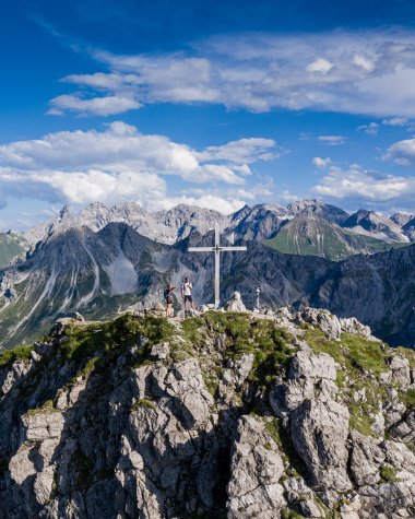 Berggipfel mit Gipfelkreuz und Wanderern, oberhalb der Alpen, bei schönem Wetter, mit blauer Himmel und Wolken. | © Kleinwalsertal Tourismus eGen • Fotograf: Bastian Morell