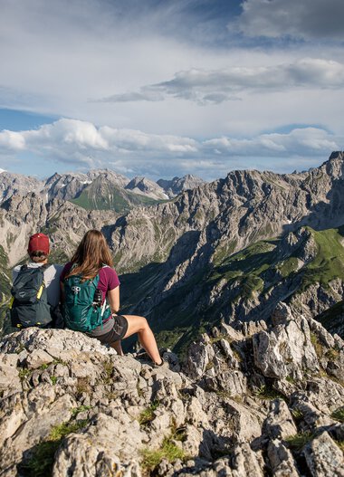 Zwei Wanderer mit Rucksäcken sitzen auf Felsen und genießen die Bergkulisse der Kanzelwand mit Gipfel im Hintergrund bei schönem Wetter. | © Kleinwalsertal Tourismus eGen • Fotograf: Bastian Morell