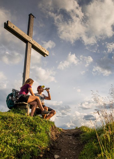 Zwei Wanderer sitzen auf einem Hügel mit einem großen Holzkruzifix im Hintergrund und genießen eine Brotzeit mit Blick auf den Himmel. | © Kleinwalsertal Tourismus eGen • Fotograf: Bastian Morell
