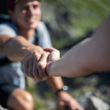 Zwei Wanderer bei einer Bergtour greifen sich herzlich die Hand, um sich gegenseitig zu unterstützen. | © Kleinwalsertal Tourismus eGen • Fotograf: Bastian Morell