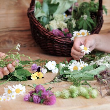 Menschen pflücken Kräuter und Blumen für ein Blumen- oder Kräutershooting an einem Holztisch, mit einem Korb voller Blumen im Hintergrund. | © Kleinwalsertal Tourismus eGen • Fotograf: Christoffer Leitner