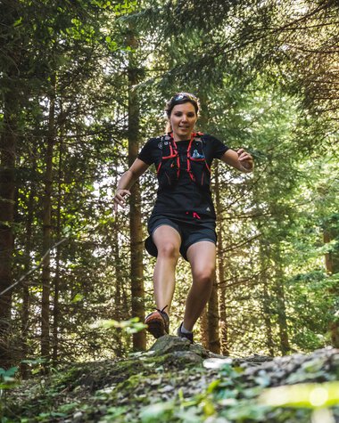 Frau beim Trailrunning in einem dichten Wald, springt fröhlich über einen Baumstamm. | © Kleinwalsertal Tourismus eGen • Fotograf: Dominik Berchtold