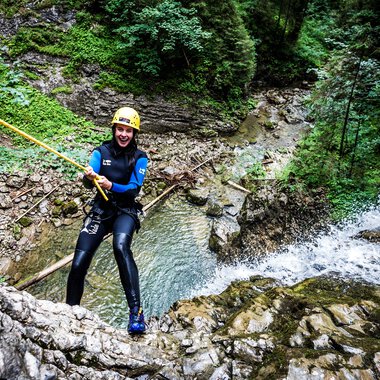 Frau beim Canyoning in einem Fluss inmitten eines grünen Waldes, trägt Neoprenanzug, Helm und ist an einem Seil befestigt. | © Kleinwalsertal Tourismus eGen • Fotograf: Dominik Berchtold