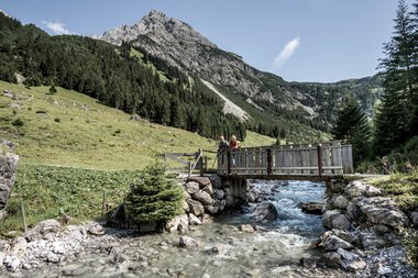 Zwei Personen auf einer Holzbrücke über einen Bach in einer alpinen Landschaft mit Bergen im Hintergrund. | © Kleinwalsertal Tourismus eGen • Fotograf: Dominik Berchtold