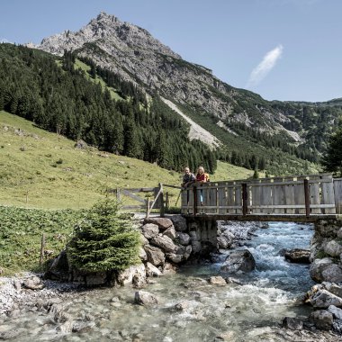 Zwei Personen auf einer Holzbrücke über einen Bach in einer alpinen Landschaft mit Bergen im Hintergrund. | © Kleinwalsertal Tourismus eGen • Fotograf: Dominik Berchtold
