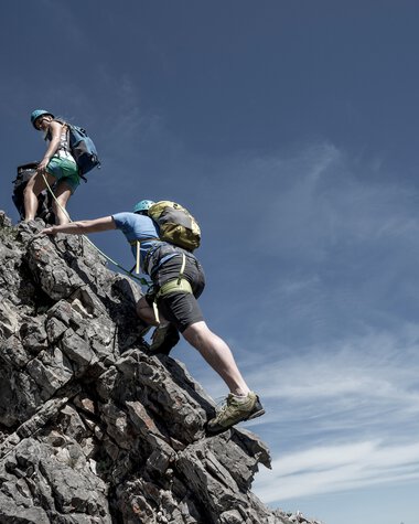Zwei Kletterer steigen eine felsige Bergspitze empor, begleitet von einem weiteren Kletterer, bei schönem Wetter und klarem Himmel. | © Kleinwalsertal Tourismus eGen • Fotograf: Dominik Berchtold
