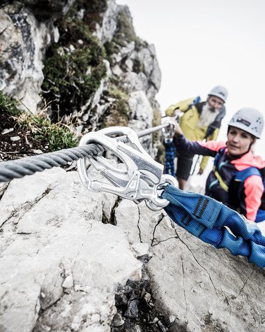 Zwei Kletterer mit Helm und Sicherheitsausrüstung beim Klettersteig an einer Felswand | © Kleinwalsertal Tourismus eGen • Fotograf: Dominik Berchtold