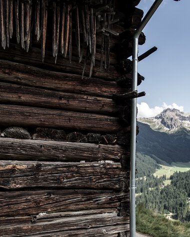 Blick auf eine Holzscheune in den Bergen mit grünen Tälern und Bergen im Hintergrund, sonniger Himmel. | © Kleinwalsertal Tourismus eGen • Fotograf: Dominik Berchtold