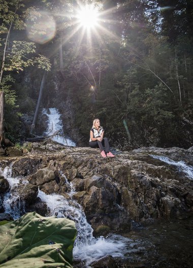 Person sitzt auf Felsen im Schwarzwasserbach in einem Wald, Sonnenlicht scheint durch die Bäume, Wasser plätschert um sie herum. | © Kleinwalsertal Tourismus eGen • Fotograf: Dominik Berchtold
