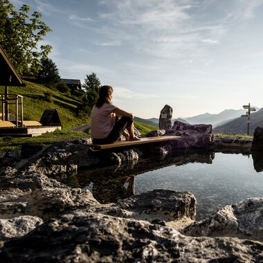 Frau sitzt am Naturerlebnisplatz Rohrweg, umgeben von Felsen, mit Blick auf Berge und Sonnenuntergang. | © Kleinwalsertal Tourismus eGen • Fotograf: Dominik Berchtold