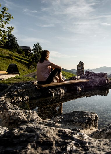 Frau sitzt am Naturerlebnisplatz Rohrweg, umgeben von Felsen, mit Blick auf Berge und Sonnenuntergang. | © Kleinwalsertal Tourismus eGen • Fotograf: Dominik Berchtold