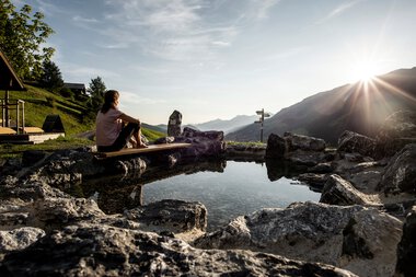 Frau sitzt am Naturerlebnisplatz Rohrweg, umgeben von Felsen, mit Blick auf Berge und Sonnenuntergang. | © Kleinwalsertal Tourismus eGen • Fotograf: Dominik Berchtold