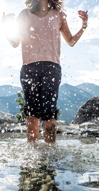 Kind läuft barfuß im Wasser an einem Naturerlebnisplatz mit Bergen im Hintergrund, Wasser spritzt beim Laufen. | © Kleinwalsertal Tourismus eGen • Fotograf: Dominik Berchtold