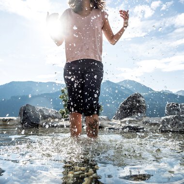 Child barefoot in water at a nature experience area, splashing as they walk with mountains in the background. | © Kleinwalsertal Tourismus eGen • Photographer: Dominik Berchtold