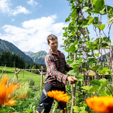 Mann in einem Permakulturgarten, umgeben von blühenden Pflanzen und grünen Bergen im Hintergrund, bei sonnigem Wetter. | © Kleinwalsertal Tourismus eGen • Fotograf: Dominik Berchtold