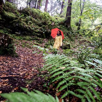 © Kleinwalsertal Tourismus eGen • Fotograf: Dominik Berchtold Frau mit rotem Regenschirm steht im Regen in einem grünen, bewaldeten Gebiet an einem bewölkten Tag. | © Kleinwalsertal Tourismus eGen • Fotograf: Dominik Berchtold