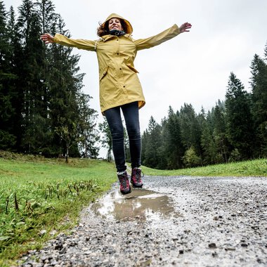 Frau in gelber Regenjacke und dunklen Hosen steht auf einer nassen Waldstraße mit Armen ausgebreitet bei schlechtem Wetter im Wald. | © Kleinwalsertal Tourismus eGen • Fotograf: Dominik Berchtold