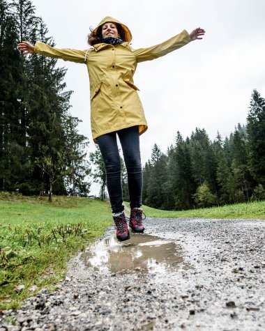 Frau in gelber Regenjacke und dunklen Hosen steht auf einer nassen Waldstraße mit Armen ausgebreitet bei schlechtem Wetter im Wald. | © Kleinwalsertal Tourismus eGen • Fotograf: Dominik Berchtold