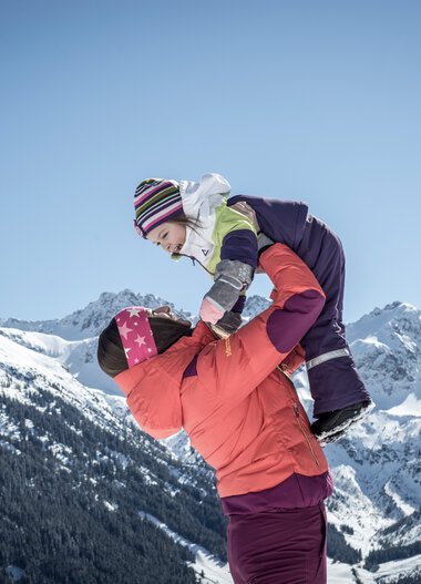 Familie beim Winterwandern in den Bergen, ein Erwachsener hebt ein Kind auf einem Schneehügel, im Hintergrund verschneite Berge unter blauem Himmel. | © Kleinwalsertal Tourismus eGen • Fotograf: Dominik Berchtold