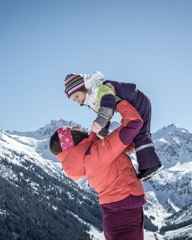 Familie beim Winterwandern in den Bergen, ein Erwachsener hebt ein Kind auf einem Schneehügel, im Hintergrund verschneite Berge unter blauem Himmel. | © Kleinwalsertal Tourismus eGen • Fotograf: Dominik Berchtold