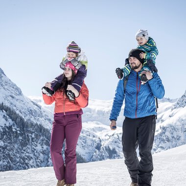 Familie beim Winterwandern in den Bergen, zwei Kinder auf den Schultern der Eltern, verschneite Gipfel im Hintergrund. | © Kleinwalsertal Tourismus eGen • Fotograf: Dominik Berchtold