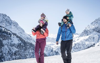 Family winter hiking in the mountains, two children on parents' shoulders, snowy peaks in the background. | © Kleinwalsertal Tourismus eGen • Photographer: Dominik Berchtold