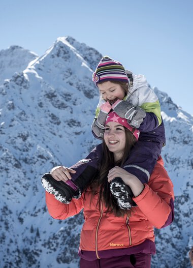 Familie beim Winterwandern in den Bergen, Kinder auf den Schultern der Eltern, mit schneebedeckten Gipfeln im Hintergrund. | © Kleinwalsertal Tourismus eGen • Fotograf: Dominik Berchtold