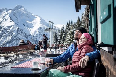 Ein Paar entspannt sich bei Kaffee im Winterurlaub in einer Berghütte mit Blick auf schneebedeckte Berge. | © Kleinwalsertal Tourismus eGen • Fotograf: Dominik Berchtold