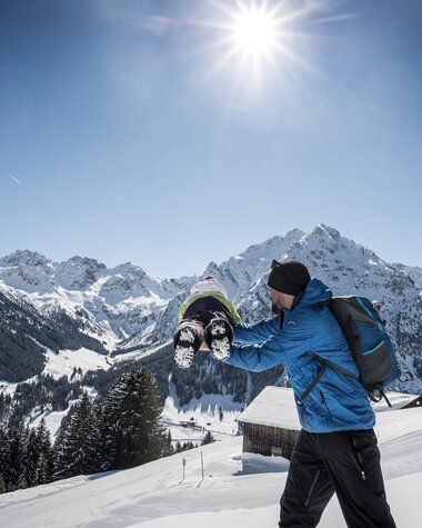 Familie beim Winterwandern in den verschneiten Bergen bei sonnigem Himmel, Vater trägt Kind auf Schultern, beeindruckende Bergkulisse im Hintergrund. | © Kleinwalsertal Tourismus eGen • Fotograf: Dominik Berchtold